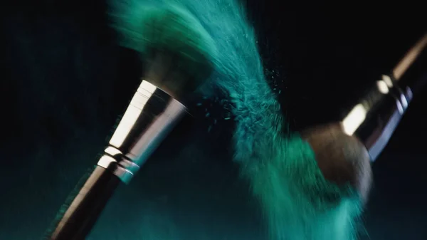Blurred cosmetic brushes with vibrant turquoise holi paint splashing on black background — Stock Photo