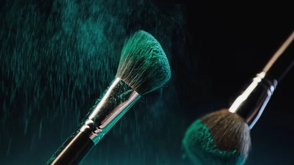 Cosmetic brushes with bright turquoise holi paint near dust splash on black background — Stock Photo