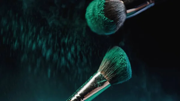 Pinceles cosméticos con pintura holi turquesa brillante cerca de salpicaduras de polvo sobre fondo oscuro - foto de stock