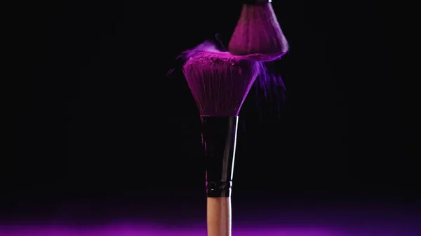 Cosmetic brushes with pink powder of holi paint hitting and splashing on black background — Stock Photo