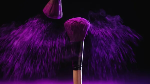 Bright cosmetic brushes with holi paint near pink dust on black background — Stock Photo