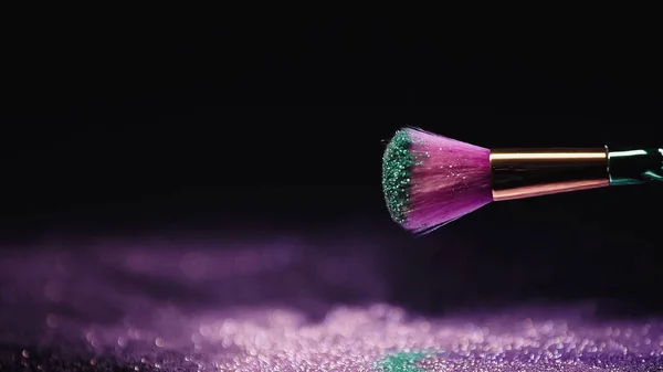 Weicher Kosmetikpinsel mit violettem und türkisfarbenem Perlstaub in der Nähe von verschwommener und glänzender Holi-Farbe auf Schwarz — Stockfoto