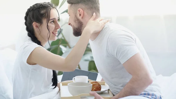 Petite amie joyeuse avec des tresses étreignant homme heureux apportant plateau de petit déjeuner au lit — Photo de stock