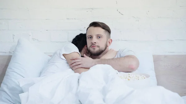 Scared woman hugging boyfriend watching scary movie near popcorn in bed — Stock Photo