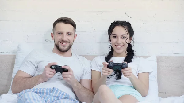 KYIV, UCRANIA - 17 de junio de 2021: joven pareja alegre jugando videojuegos en el dormitorio - foto de stock