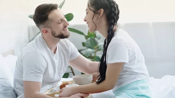 Happy brunette woman with braids bringing breakfast tray to boyfriend in bed — Stock Photo