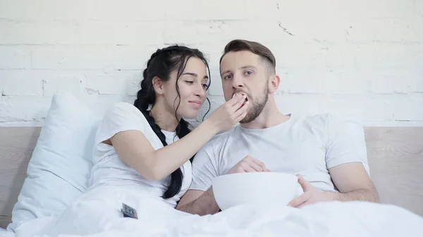 Happy young couple eating popcorn and watching movie in bed — Stock Photo