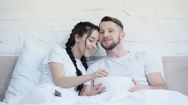 Feliz jovem casal com pipoca sorrindo e assistindo filme na cama — Fotografia de Stock