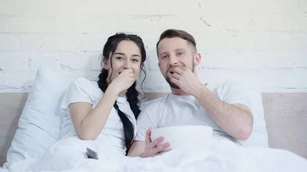 Cheerful young couple eating popcorn and watching movie in bed — Stock Photo