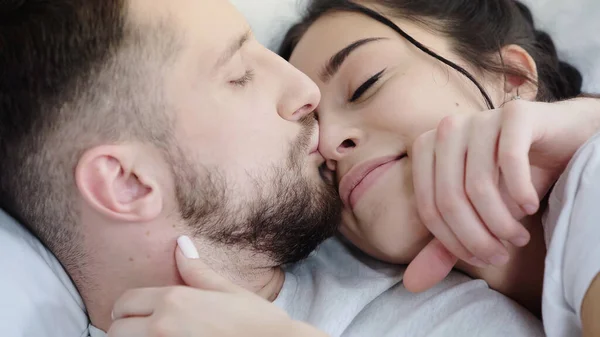 Homem barbudo beijando nariz de mulher morena feliz enquanto descansa na cama — Fotografia de Stock