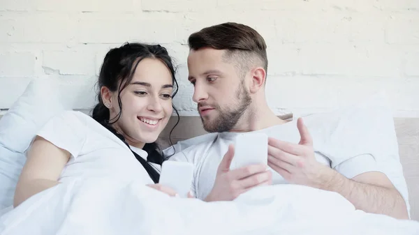 Hombre barbudo y mujer feliz sosteniendo los teléfonos celulares en casa - foto de stock