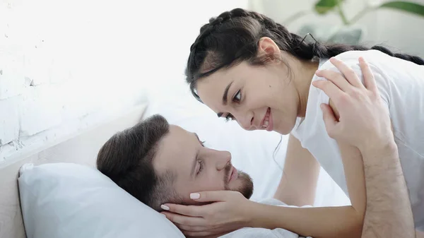 Улыбающаяся молодая женщина трогает лицо бородатого мужчины в спальне — стоковое фото