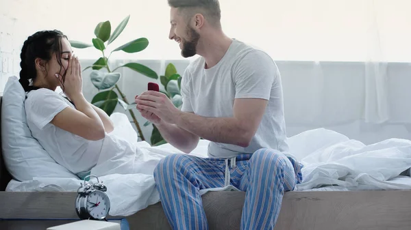 Happy bearded man holding red box with wedding ring while doing proposal to astonished girlfriend in bedroom — Stock Photo