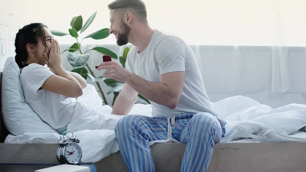 Happy bearded man holding red box with wedding ring while doing proposal to girlfriend in bedroom — Photo de stock