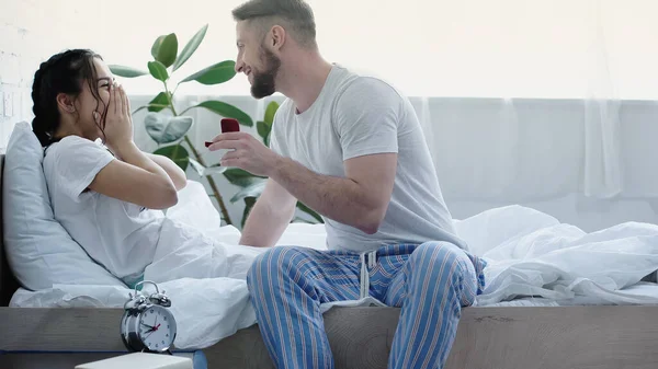 Happy bearded man holding red box with wedding ring while doing proposal to girlfriend in bedroom — Stock Photo