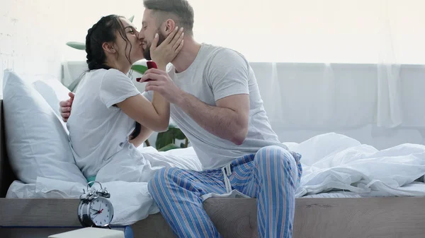 Happy bearded man holding red box with wedding ring and kissing girlfriend in bedroom — Photo de stock