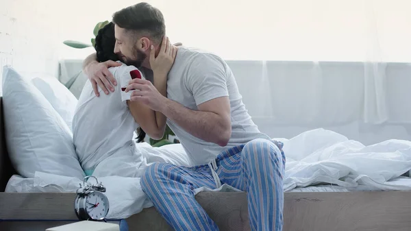 Hombre barbudo feliz sosteniendo caja roja con anillo de boda mientras abraza a la novia en el dormitorio - foto de stock