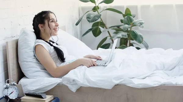 Brünette Freiberuflerin lächelt, während sie Laptop im Bett benutzt — Stockfoto