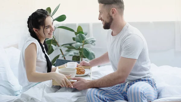 Happy man bringing breakfast tray to smiling girlfriend with braids in bedroom — Stock Photo