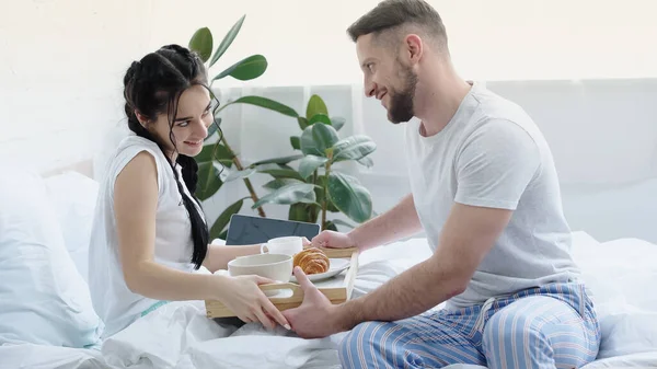 Happy man bringing breakfast tray to joyful girlfriend with braids in bedroom — Stock Photo