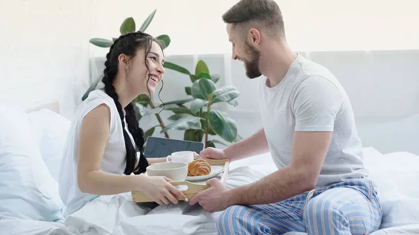 Довольный мужчина приносит завтрак улыбающейся подруге с брейками в спальне — стоковое фото