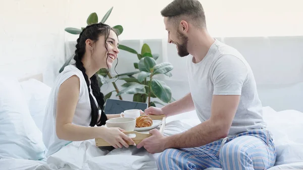 Pleased man bringing breakfast tray to smiling girlfriend with braids in bedroom — Stock Photo