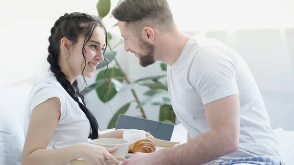 Happy man bringing breakfast to smiling girlfriend with braids in bed — Photo de stock