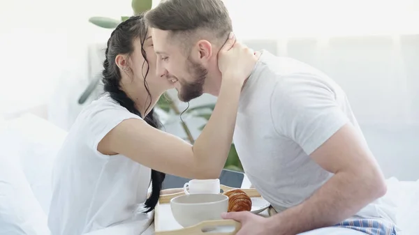 Жінка з косами цілує бородатого чоловіка, що приносить сніданок у ліжку — стокове фото