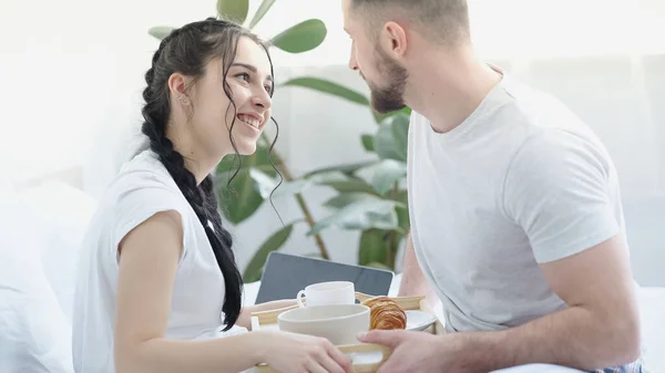 Uomo sorridente che porta la colazione alla fidanzata felice con le trecce a letto — Foto stock