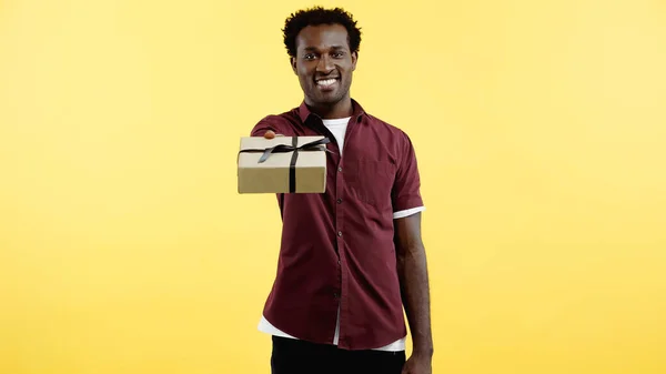 Щасливий афроамериканський чоловік у сорочці тримає загорнутий подарунок ізольовано на жовтому — стокове фото