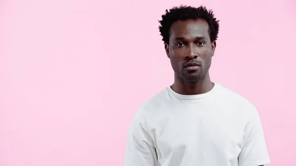 Hombre afroamericano serio en camiseta blanca mirando a la cámara aislada en rosa - foto de stock