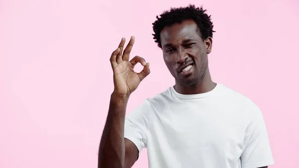 Curly african american man in white t-shirt winking eye and showing ok sign isolated on pink — Stock Photo