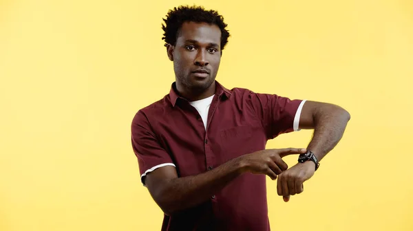 Lockiger afrikanisch-amerikanischer Mann in weinrotem Hemd zeigt auf Armbanduhr isoliert auf gelb — Stockfoto