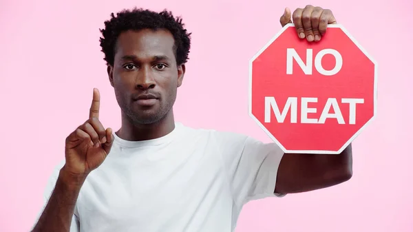 Hombre rizado afroamericano y vegetariano en camiseta blanca sosteniendo un cartel sin letras de carne y señalando con el dedo aislado en rosa - foto de stock
