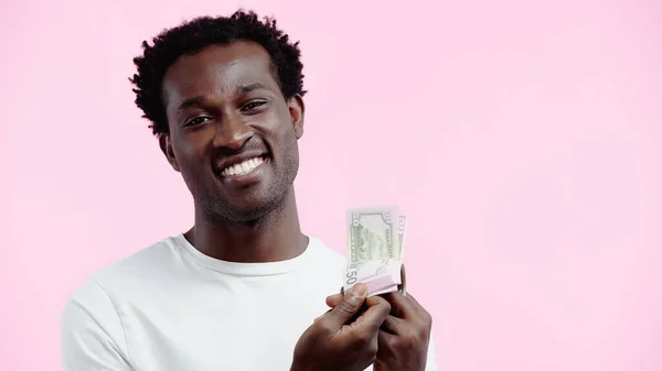 Homem americano africano alegre na t-shirt branca que olha para a câmera ao prender dólares isolados no rosa — Fotografia de Stock