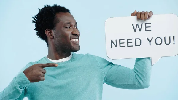Positive african american man pointing at speech bubble with we need you lettering isolated on blue — Stock Photo