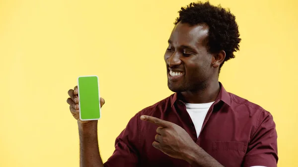 Homem americano africano feliz em camisa vermelha apontando com o dedo para o smartphone com tela verde isolada no amarelo — Fotografia de Stock