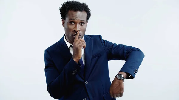 Hombre de negocios afroamericano en traje con corbata mostrando un signo de silencio mientras muestra un reloj de pulsera aislado en azul - foto de stock