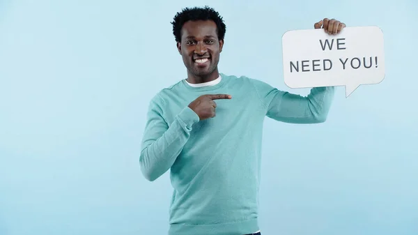 Pleased african american man pointing with finger at speech bubble with we need you lettering isolated on blue — Stock Photo