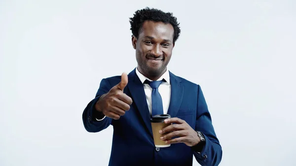 Pleased african american businessman holding paper cup and showing thumb up isolated on blue — Stock Photo