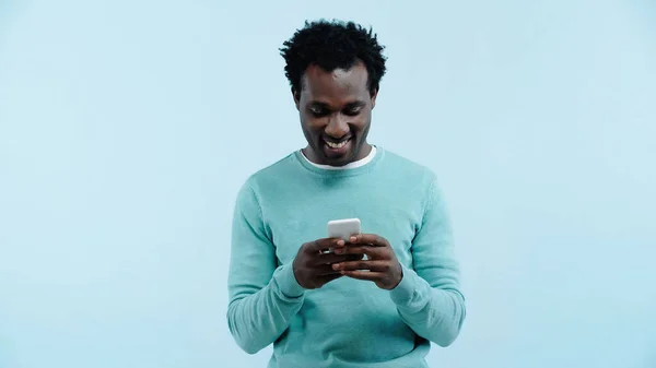 Улыбающийся африканский американец, обменивающийся сообщениями по мобильному, изолированный на голубом — стоковое фото