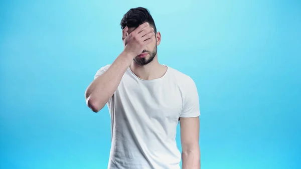 Hombre deprimido en camiseta blanca que cubre los ojos con la mano aislada en azul - foto de stock