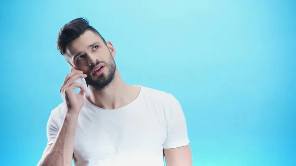 Hombre pensativo mirando hacia arriba mientras habla en el teléfono inteligente aislado en azul — Stock Photo