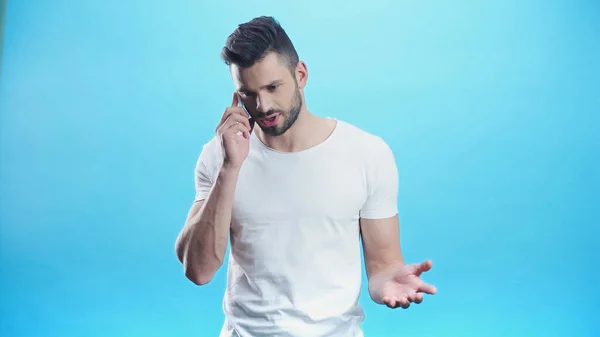 Upset man gesturing during conversation on cellphone isolated on blue — Stock Photo