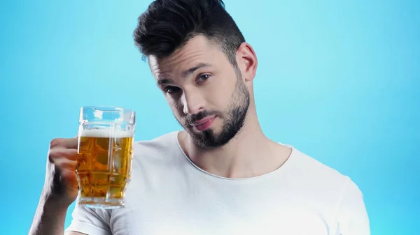 Young man looking at camera while holding mug of beer isolated on blue — Stock Photo