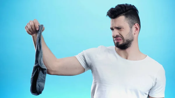 Displeased man grimacing while holding smelly sock isolated on blue — Stock Photo