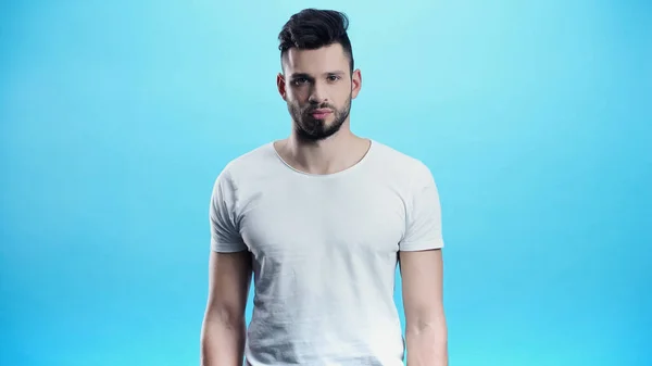 Jeune homme sérieux en t-shirt blanc regardant la caméra isolée sur bleu — Photo de stock