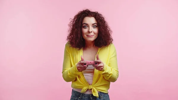 KYIV, UCRANIA - 30 de junio de 2021: una joven rizada sosteniendo el joystick y jugando un videojuego aislado en rosa - foto de stock