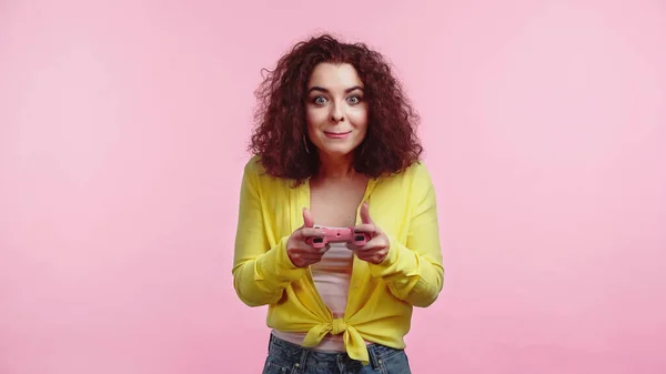 KYIV, UKRAINE - JUNE 30, 2021: young curly woman holding joystick and playing video game isolated on pink — Stock Photo