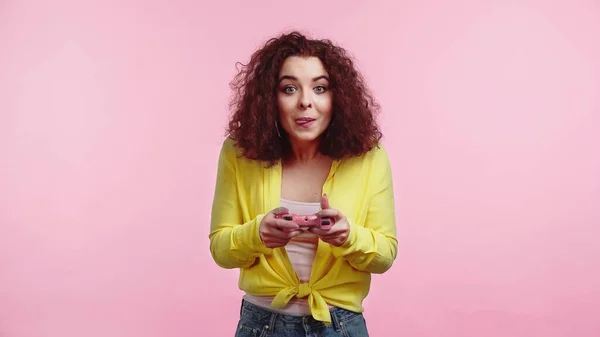 KYIV, UKRAINE - JUNE 30, 2021: young curly woman sticking out tongue and playing video game isolated on pink — Stock Photo